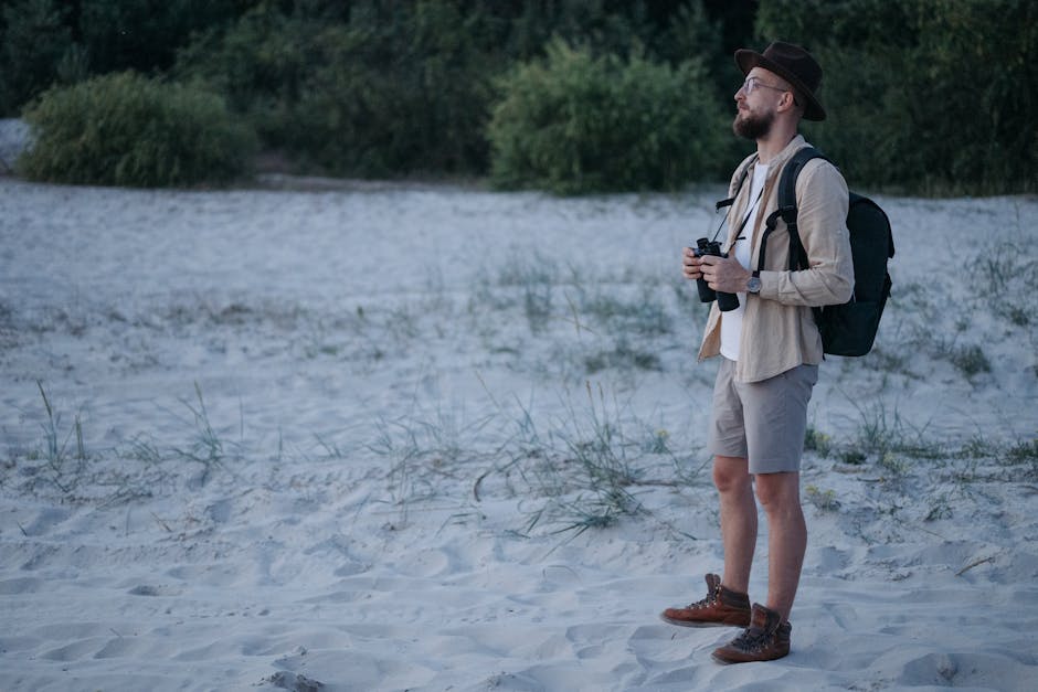 Bearded man with backpack and binoculars exploring a sandy beach.