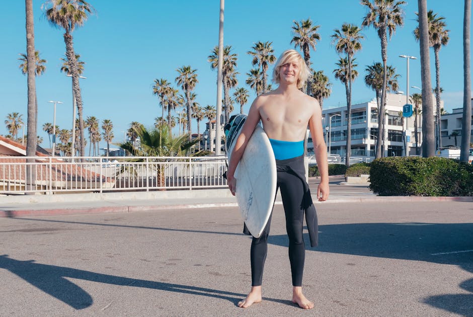 Smiling young surfer carrying a surfboard barefoot on a sunny street lined with palm trees.