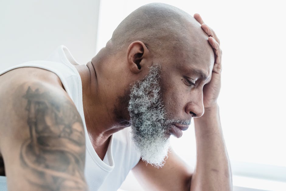Profile of a bald, bearded man deep in thought, hand on forehead, tattoo visible.