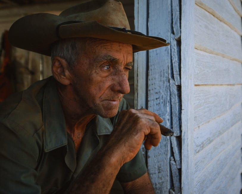 Elderly man in cowboy hat pensively gazing from a wooden porch, rustic setting.