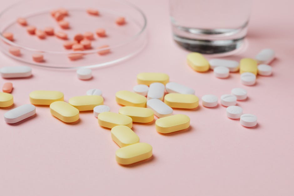A close-up of various pills and tablets on a pink background with a glass of water.