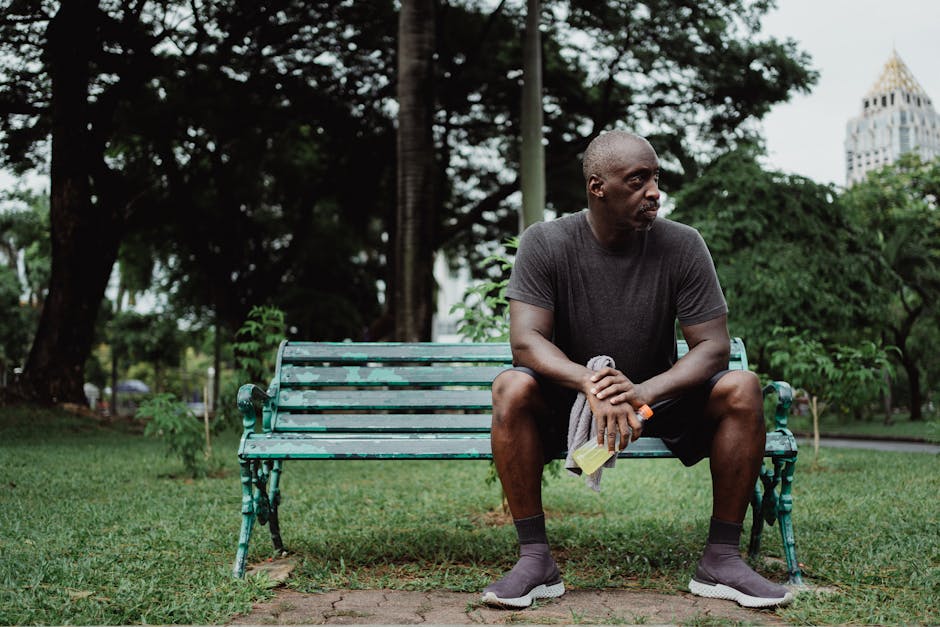 A man sits on a park bench holding a beverage bottle, appearing pensive.