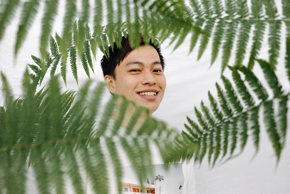 A smiling man photographed through lush green ferns, creating a vibrant, happy portrait in nature.