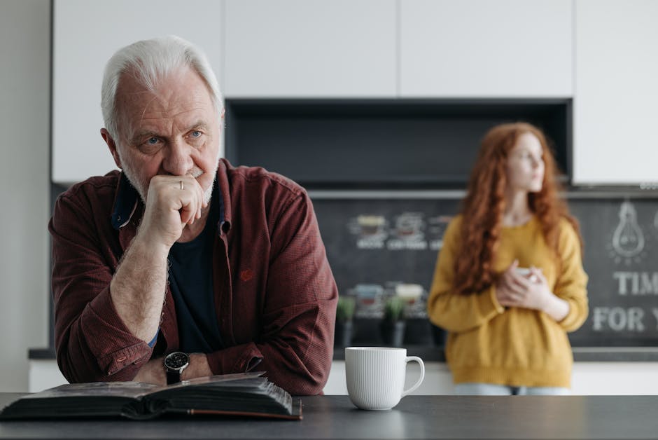 Elderly man looking thoughtful in a kitchen, with a young woman in the background, conveying a scene of contemplation and emotion.