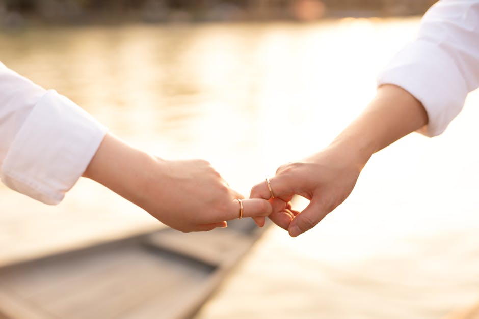 A tender moment between a couple touching hands by the water, showcasing wedding rings in soft sunlight.