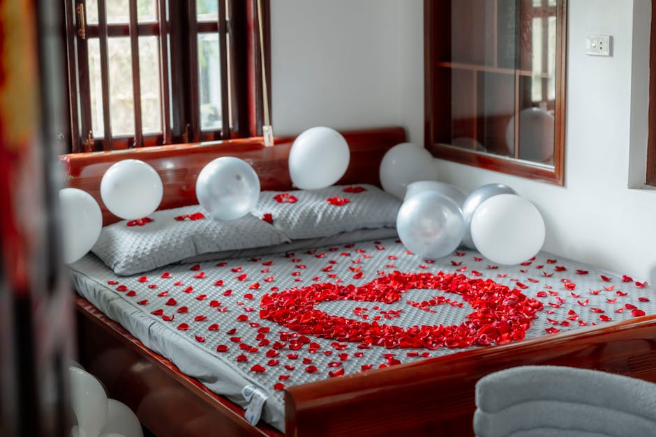 A cozy bed decorated with heart-shaped red rose petals and white balloons for a romantic setting.